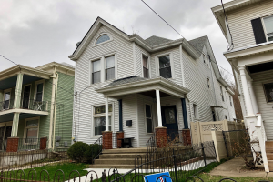 Reihe von Häusern mit Fenstern, Türen, Säulen, Geländern, Stufen und Dächern, umgeben von einem Metallzaun, Gras, Pflanzen, Bäumen und einem bewölkten Himmel, mit einem Mietschild am Boden, das die Adresse und die Details der Anzeige anzeigt.