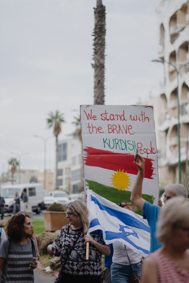 Eine Gruppe von Menschen marschiert nachts eine Straße entlang, hält ein Schild hoch, auf dem steht "Wir stehen zu den tapferen kurdischen Menschen", mit einem Van, der am Straßenrand geparkt ist, Straßenlaternen, Bäumen und Gebäuden im Hintergrund.