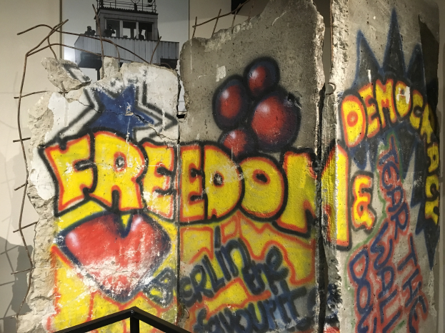 Eine Wand mit großer, schwarzer Graffiti-Aufschrift "Freiheit und Demokratie", einem Geländer unten und einem Gebäude mit Fenstern im Hintergrund.