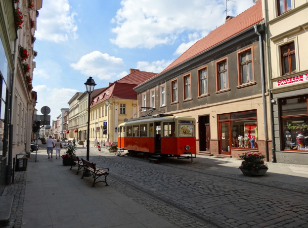 Eine rote und gelbe Straßenbahn fährt durch die Kopfsteinpflasterstraße der Altstadt von Tallinn, flankiert von Gebäuden mit Fenstern, Bänken und Laternen, mit Menschen auf den Bürgersteigen und einem bewölkten Himmel im Hintergrund.
