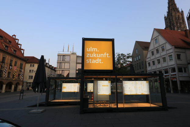 Ein Bushaltestelle in einem Stadtplatz mit einem geparkten Auto davor, umgeben von Gebäuden, Bäumen und Fahnenmasten, mit einer Tafel im Vordergrund und einem klaren blauen Himmel im Hintergrund.