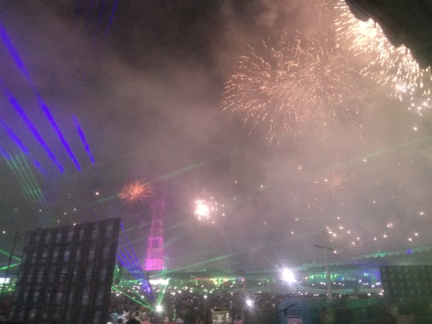 Eine Menschenmenge bei einem Konzert, beleuchtet von Gebäuden und Laternen, mit farbigen Feuerwerken am Himmel.