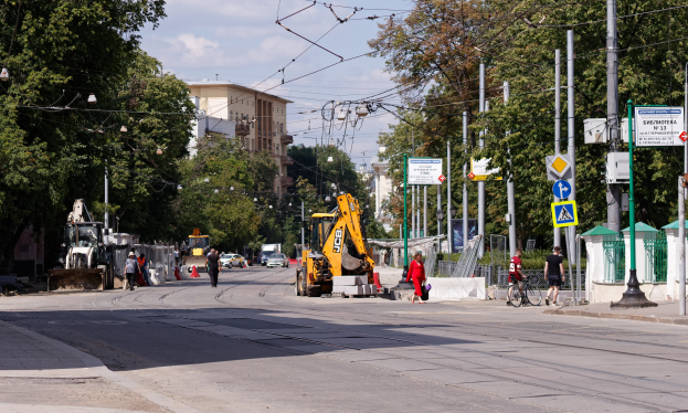 Eine Stadtstraße mit einer Baustelle, Fahrzeugen, Fußgängern, einem Radfahrer, Verkehrskegeln, Pfählen, Schildern, Strommasten mit Drähten, Bäumen, Gebäuden mit Fenstern und einem bewölkten Himmel.