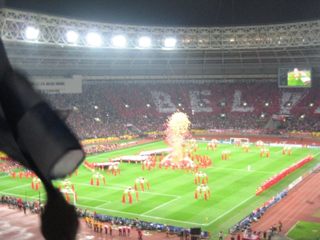 Ein Stadion mit Flutlicht, Eisengittern, Zuschauern, stehenden Personen, Werbetafeln, Bildschirmen, einer Überwachungskamera und Fahnen und Dekorationen.