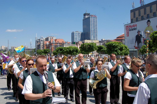 Eine Gruppe von Menschen in einheitlichen Kostümen, die verschiedene Musikinstrumente spielen, vor Bäumen, Pfählen und Gebäuden, mit einer Banner mit Text und Bildern auf der rechten Seite.