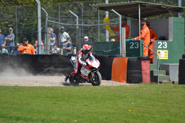 Eine Person in Helm und Handschuhen auf einem Motorrad auf einem grasbewachsenen Feld mit Zuschauern, Absperrung, Treppe und einem Schuppen im Hintergrund.