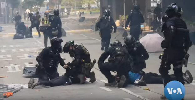 Eine Gruppe von Polizisten in Einsatzausrüstung umgibt einen Mann, der auf dem Boden liegt, mit Bäumen, Masten, Gebäuden und anderen Gegenständen im Hintergrund.
