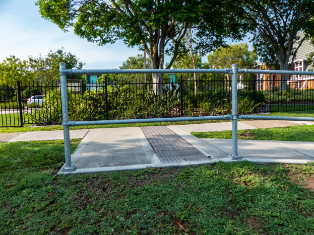 Ein Metallzaun in einer grasigen Fläche, umgeben von Pflanzen, Bäumen und einem Auto auf der Straße, mit Gebäuden und einem klaren blauen Himmel im Hintergrund.