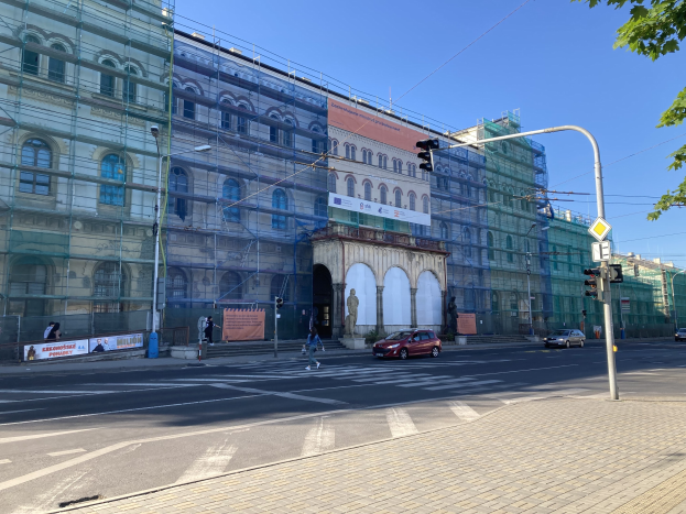 Ein großes Gebäude mit Gerüst, umgeben von Fahrzeugen, Fußgängern und städtischer Infrastruktur unter einem klaren blauen Himmel.