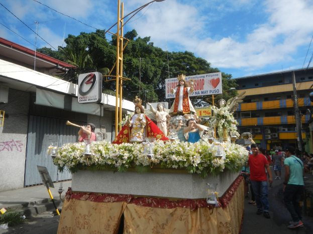 Ein Paradewagen mit Statuen, Blumen und Texttafeln, der durch eine Straße mit Laternenmasten, Gebäuden, Bäumen und einem bewölkten Himmel fährt.