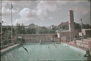 Altes Schwarz-Weiß-Foto von Schwimmern in einem Pool mit Gebäuden, Bäumen, Pfählen, Fahnen und einem Uhrenturm im Hintergrund unter einem bewölkten Himmel.
