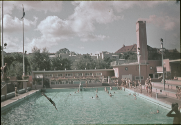 Altes Schwarz-Weiß-Foto von Schwimmern in einem Pool mit Gebäuden, Bäumen, Pfählen, Fahnen und einem Uhrenturm im Hintergrund unter einem bewölkten Himmel.