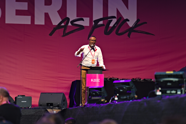 Ein Mann in einem weißen Hemd und braunen Hosen steht auf einer Bühne und hält ein Mikrofon in der Hand, mit einem pinken Banner dahinter, das 'Berlin as Fuck' lautet, und Lautsprechern auf der Bühne.