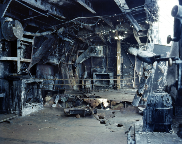 Verkohlte Trümmer und verstreute Metallobjekte in einem feuerbeschädigten Gebäude mit sichtbarem Himmel durch die Decke.