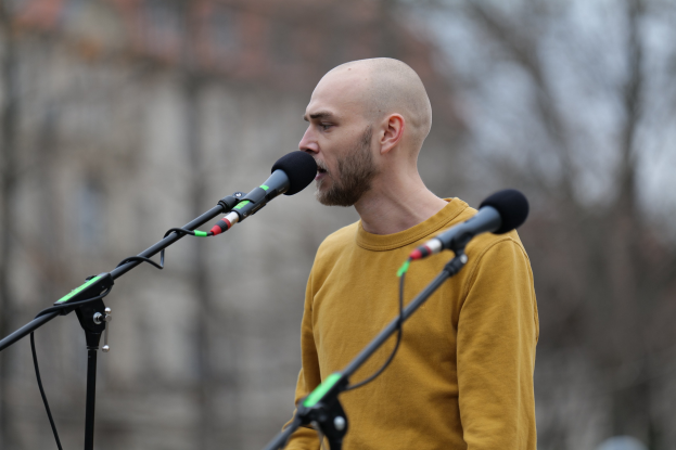 Ein kahlkopfiger Mann in einem gelben T-Shirt singt in ein Mikrofon gegen einen unscharfen Hintergrund.