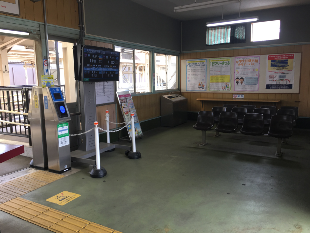 Ein belebter Bahnhofseingang mit Sitzgelegenheiten, einer Informations counter, Ticketautomaten, Kettenpollern, Informationsanzeigen, Wandplakaten, Deckenbeleuchtung und Geländern.