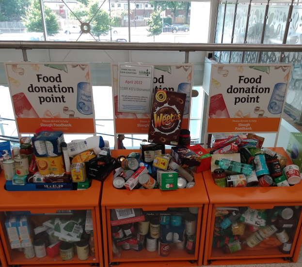 Orange Schübe mit Lebensmittelspenden gefüllt, darunter Flaschen, Dosen und Kisten, mit Informationsschildern und Blick durch Glasfenster auf Fahrzeuge, Bäume, Gebäude und Himmel.