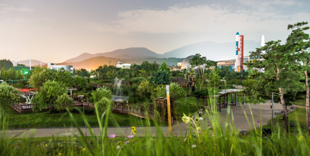 Ein Park mit grünem Gras, bunten Blumen, hohen Bäumen, einer Brücke, einem Springbrunnen, Laternenmasten und fernen Gebäuden vor Bergen und einem klaren blauen Himmel.