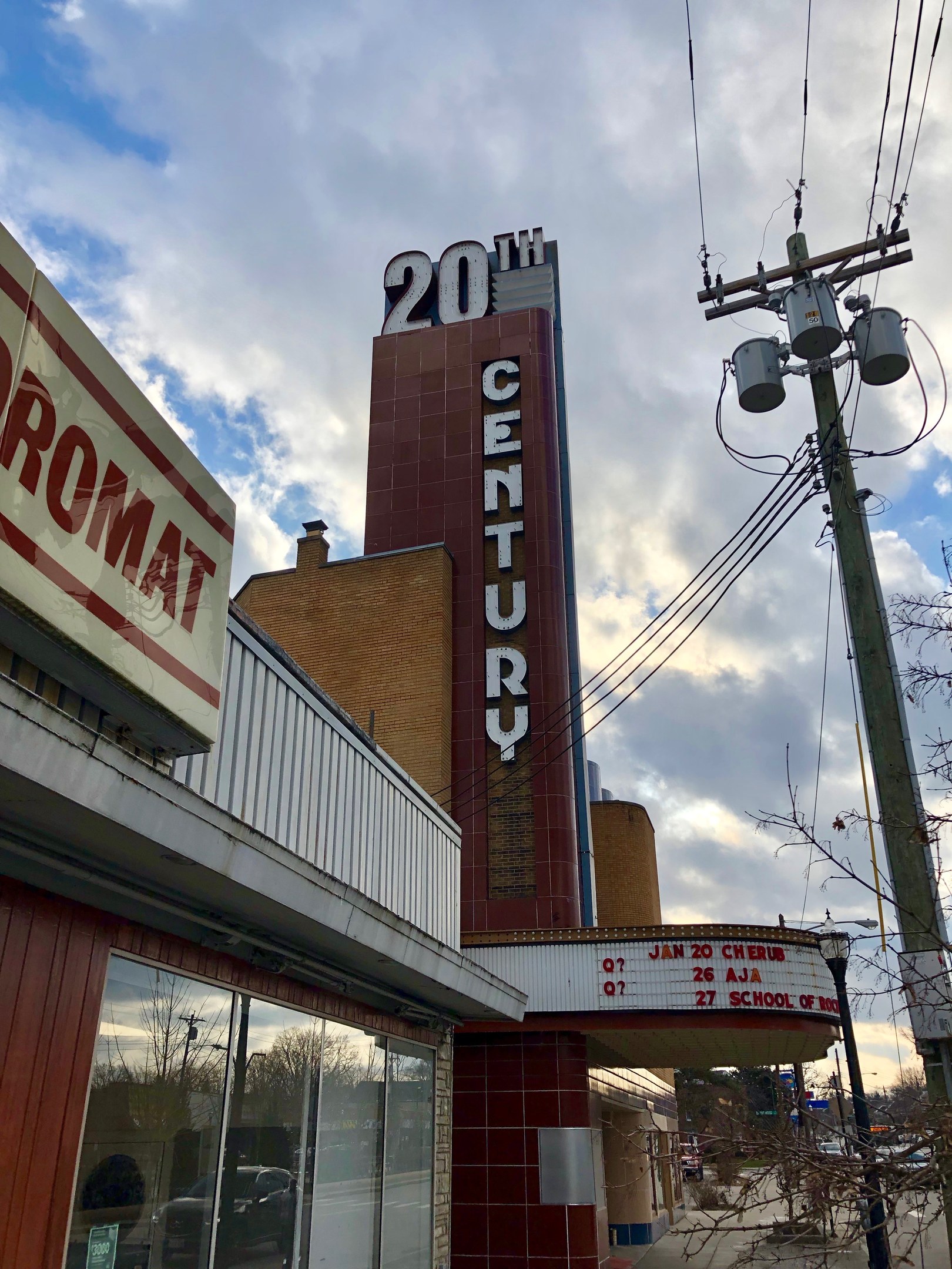 Außenansicht des 20th Century Theatre in Detroit, Michigan, mit Glaswänden und Namensschildern, umgeben von Bäumen und Strommasten, mit einem bewölkten Himmel im Hintergrund.