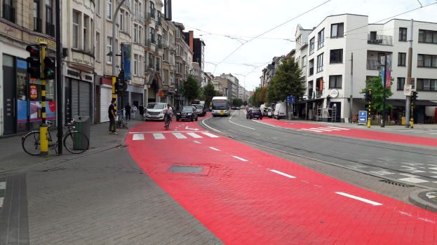 Eine Stadtstraße mit einem roten Fahrradweg, Fahrzeugen, Fußgängern, Gebäuden, Bäumen und einem klaren blauen Himmel.