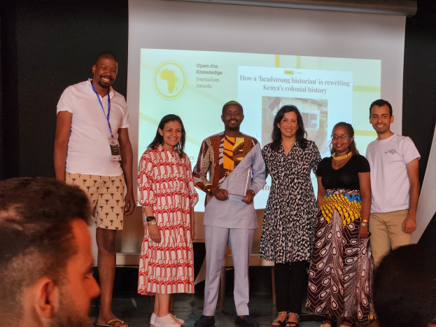 Eine Gruppe von Menschen auf einer Bühne, die lächeln und für ein Foto posieren, mit einem Publikum darunter und einem Bildschirm im Hintergrund, der "Open the Knowledge Journalism Awards 2019" anzeigt.