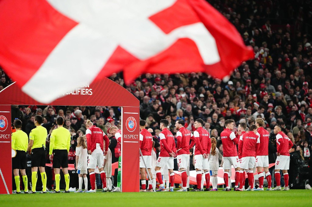 Eine Gruppe von Menschen auf einem Fußballfeld mit einer roten und weißen Flagge im Vordergrund, einem Bogen mit Text "Bayern Munich vs Bayern Munich Betting Tips & Preview" im Hintergrund und einer großen Menge im Stadion.