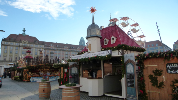 Weihnachtsmarkt auf einem Stadtplatz mit geschmückten Bäumen, Gebäuden, Fahrzeugen, Menschen, Straßenlaternen und einem bewölkten Himmel.