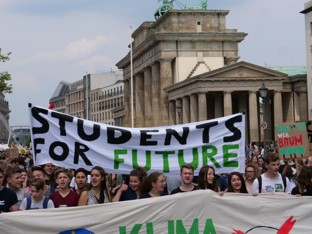 Eine Gruppe von Schülern marschiert in Berlin, eine bunt bemalte Fahne mit der Aufschrift "Schüler für die Zukunft" schwingend, mit Gebäuden, Bäumen und Himmel im Hintergrund.
