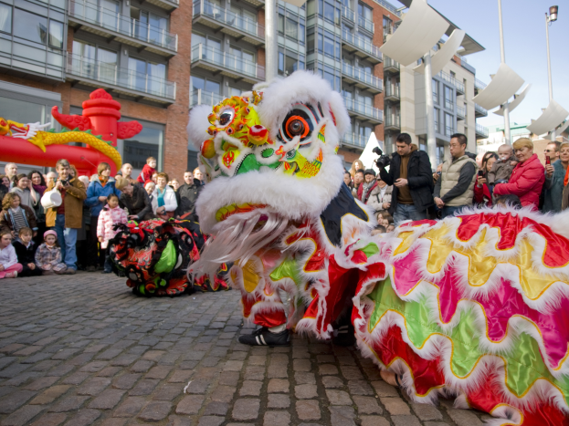 Eine Löwentanzvorstellung während eines Chinesischen Neujahrsfests in Amsterdam, mit einer Menge Menschen, die zuschauen und den Moment mit Kameras festhalten, vor einem Hintergrund von Gebäuden und einem klaren blauen Himmel.