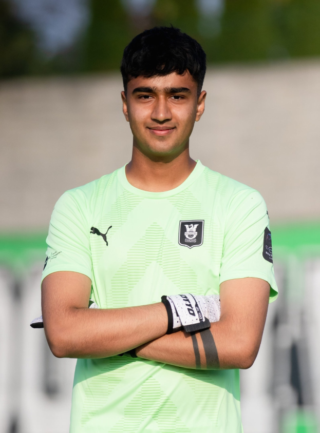 Ein junger Mann in einem grünen Hemd steht auf einem Fußballfeld mit verschränkten Armen, strahlend, mit einem unscharfen Hintergrund.