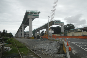 Baustelle mit einer Brücke im Hintergrund, Straße mit Absperrgittern auf der rechten Seite, Bahnschiene auf der linken Seite, Steine und Gras am Boden, Bäume und Gebäude auf beiden Seiten der Straße und ein bewölkter Himmel.
