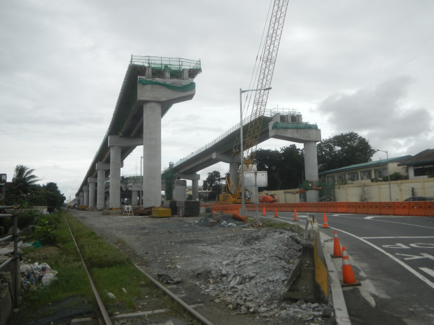 Baustelle mit einer Brücke im Hintergrund, Straße mit Absperrgittern auf der rechten Seite, Bahnschiene auf der linken Seite, Steine und Gras am Boden, Bäume und Gebäude auf beiden Seiten der Straße und ein bewölkter Himmel.