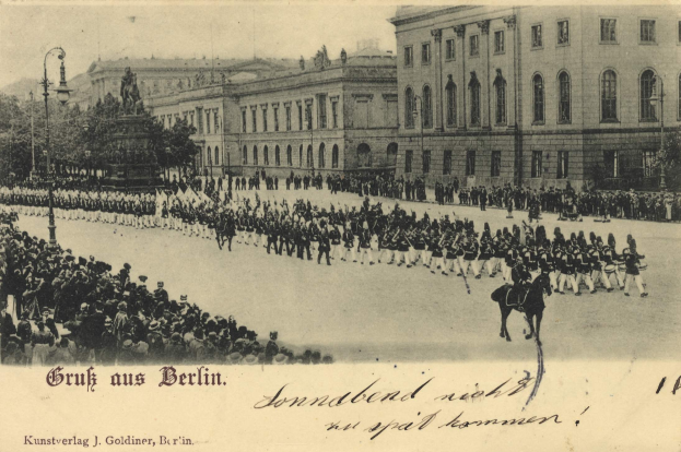 Ein altes Schwarz-Weiß-Foto einer Parade in Berlin, Deutschland, mit Menschen auf dem Boden und auf Pferden, einer Statue, Straßenlaternen, Bäumen, Gebäuden mit Fenstern und einem bewölkten Himmel, mit Text unten.