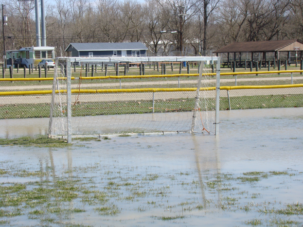 Ein Fußballtor steht inmitten eines überfluteten Feldes, umgeben von Gras und Wasser, mit Hütten, Pfosten, Bäumen, Fahrzeugen und einem klaren blauen Himmel im Hintergrund.