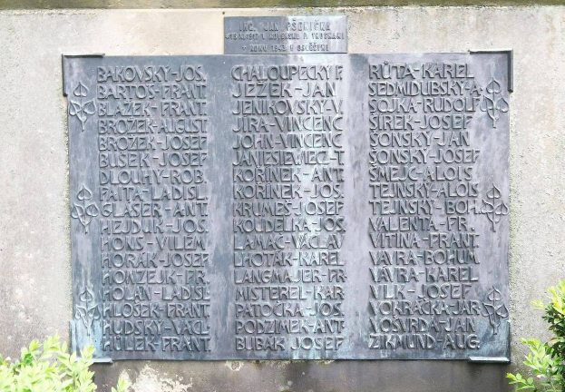 Ein Steinplakette an einer Wand mit eingraviertem Text, umgeben von Pflanzen am Boden, wahrscheinlich ein Denkmal für Holocaust-Opfer.