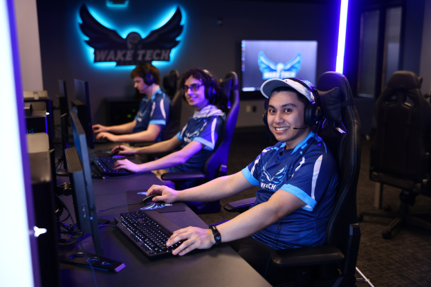 Gruppe von Menschen mit Headsets, die während eines Esports-Turniers lächeln und an Computern sitzen, mit Monitoren, Tastaturen und Stühlen im Bild.