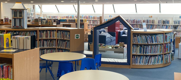 Ein Bibliotheksinneres mit Regalen, Tischen, St├╝hlen, Sofas mit Kissen, einer Stange, Deckenleuchten und anderen Gegenst├Ąnden.