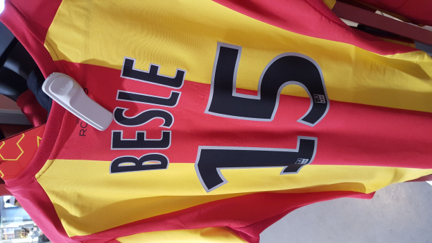 Rotes und gelbes Fußballtrikot mit Text und Nummern auf einem Ladengestell hängend, mit anderen Gegenständen und dem Boden im Hintergrund.