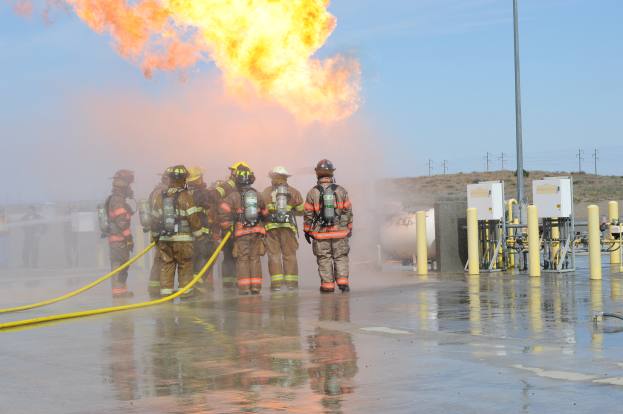 Eine Gruppe von Feuerwehrleuten in Helmen mit Rohren vor einem großen Feuer, mit Pfählen, Drähten und anderen Gegenständen auf dem Boden und einem bewölkten Himmel im Hintergrund.