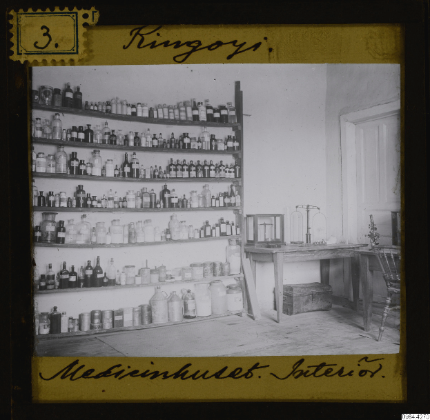 Ein altes Schwarz-Weiß-Foto eines Apothekeninneren mit Regalen voller Flaschen, einem Tisch mit verschiedenen Gegenständen, einem Stuhl, einer Tür und einer Wand, mit Text am unteren Bildrand.