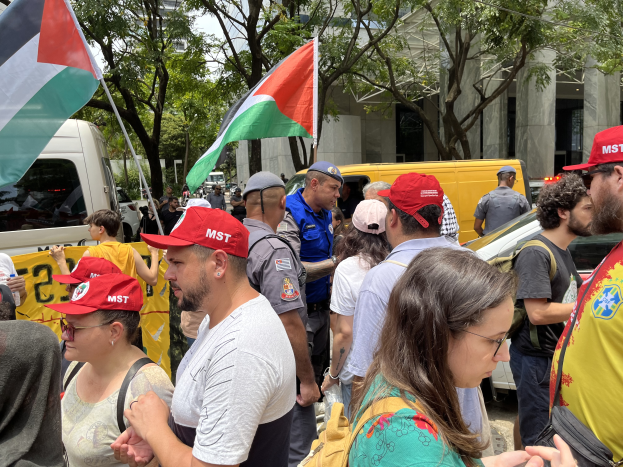 Eine Gruppe von Menschen steht vor einem Gebäude, einige tragen Mützen und tragen Taschen, halten palästinensische Flaggen, mit Fahrzeugen auf der Straße, Bäumen im Hintergrund und einem klaren blauen Himmel.