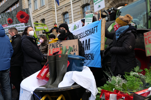 Maskierte Menschen mit Protestschildern vor einem Lkw, mit einem Tisch mit verschiedenen Gegenständen, Topfpflanzen und einem beflaggten Gebäude im Hintergrund.