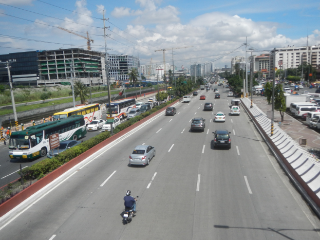 Busy city street with cars, buses, motorcycles, trees, buildings, poles, wires, and a cloudy sky.