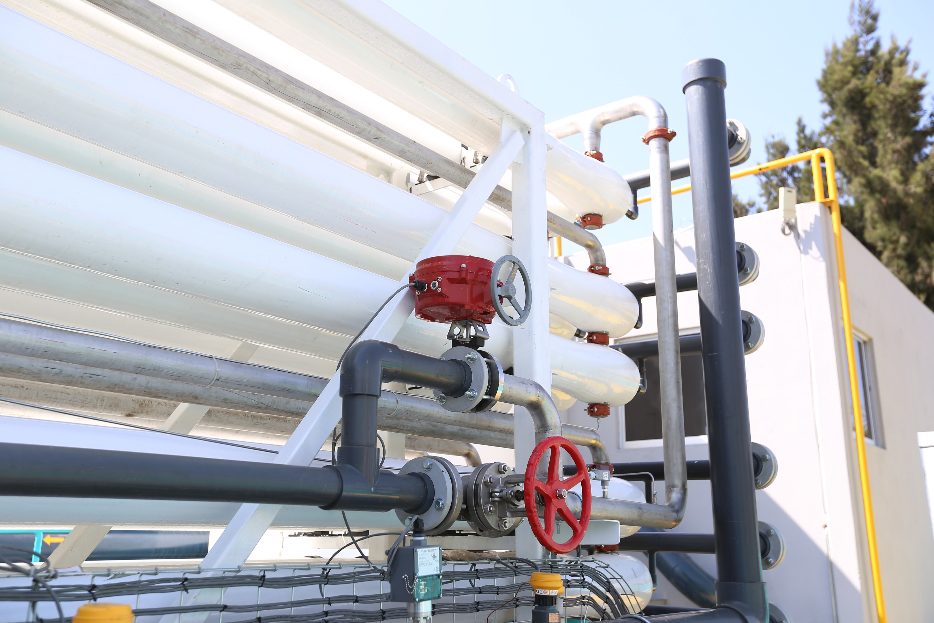 Große Gruppe von Rohren, Ventilen, Metallstangen und anderen Gegenständen auf einem Gebäudedach mit Bäumen und einem klaren blauen Himmel im Hintergrund.