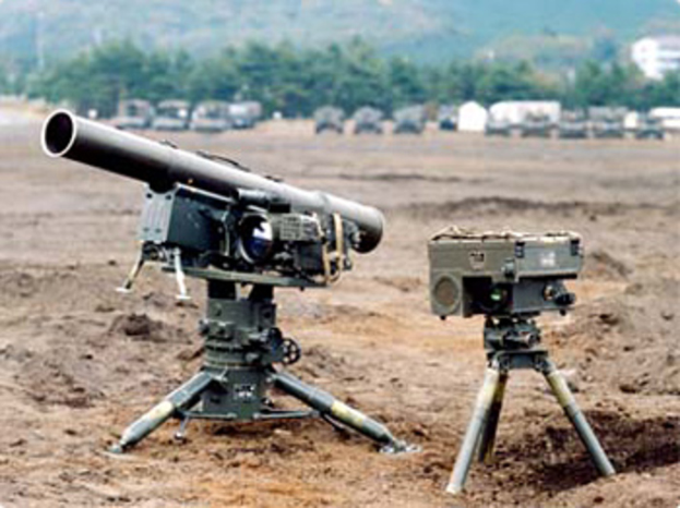 Ein Raketensystem ist auf dem Boden angeordnet mit im Hintergrund geparkten Fahrzeugen, sowie Bäumen, einem Gebäude und einem Hügel.