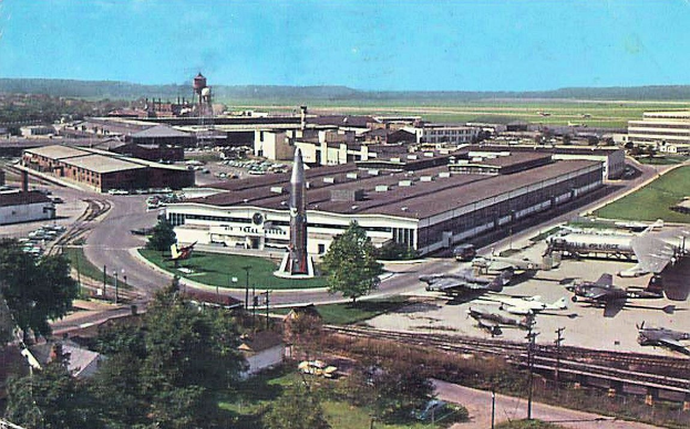 Altes Schwarz-Weiß-Foto eines Flughafens mit parkenden Flugzeugen, Gebäuden, Bäumen, Straßen, Fahrzeugen, Strommasten, Gras, Bahnschienen, Türmen, Hügeln und einem klaren Himmel, mit Text unten.