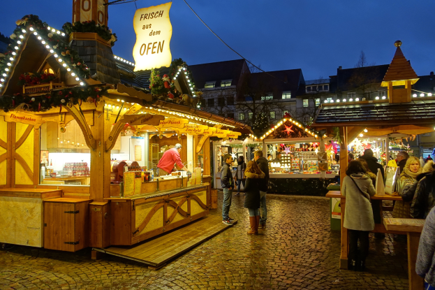 Ein lebendiger Weihnachtsmarkt auf einem Kopfsteinpflasterweg bei Nacht, mit Menschen, die an Ständen mit Lichtern und festlichen Dekorationen vorbeischlendern, Gebäuden, Bäumen und einem klaren Nachthimmel im Hintergrund.