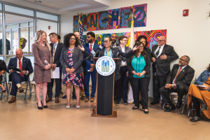 Eine Gruppe von Menschen steht vor einem Podium mit einem Mikrofon und einer Tafel mit dem Text "Sancho-Stadtrat tritt dem Stadtrat bei", während andere auf Stühlen sitzen oder in der Nähe stehen; Flaggen, ein Feuerlöscher und Deckenleuchten sind an der hinteren Wand zu sehen.