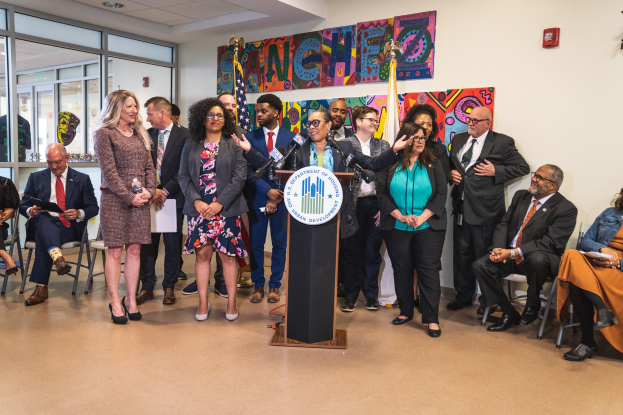 Eine Gruppe von Menschen steht vor einem Podium mit einem Mikrofon und einer Tafel mit dem Text "Sancho-Stadtrat tritt dem Stadtrat bei", während andere auf Stühlen sitzen oder in der Nähe stehen; Flaggen, ein Feuerlöscher und Deckenleuchten sind an der hinteren Wand zu sehen.