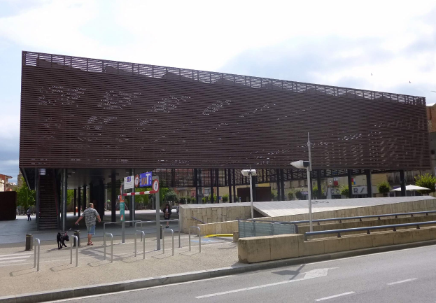 Großes mehrstöckiges Gebäude mit zahlreichen Fenstern, das als neuer Eingang zum Stadtzentrum dient, flankiert von Straßeninfrastruktur wie Pfählen, Laternen und Schildern, mit Fußgängern, Fahrzeugen, Bäumen und bewölktem Himmel im Hintergrund.